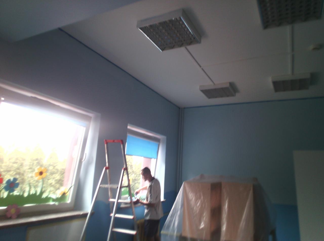 Mężczyzna maluje ścianę w pomieszczeniu z oknami ozdobionymi kwiatami, drabina oparta o ścianę, meble zabezpieczone folią, widoczne lampy sufitowe.