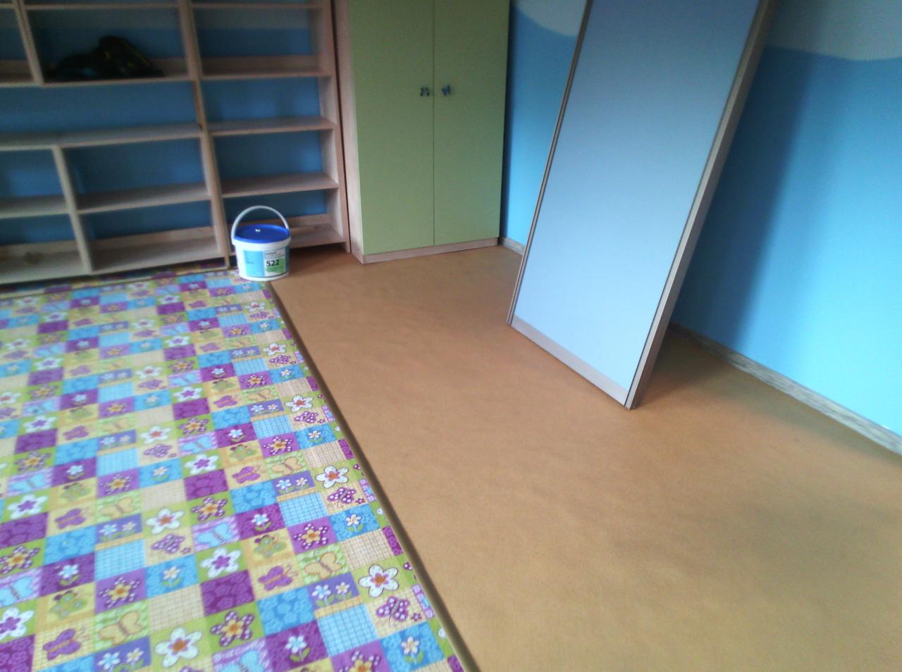 Podłoga wyłożona płytami OSB, malowana ściana w kolorze błękitnym, regał z półkami, zielona szafka i wiadro z farbą w pokoju.