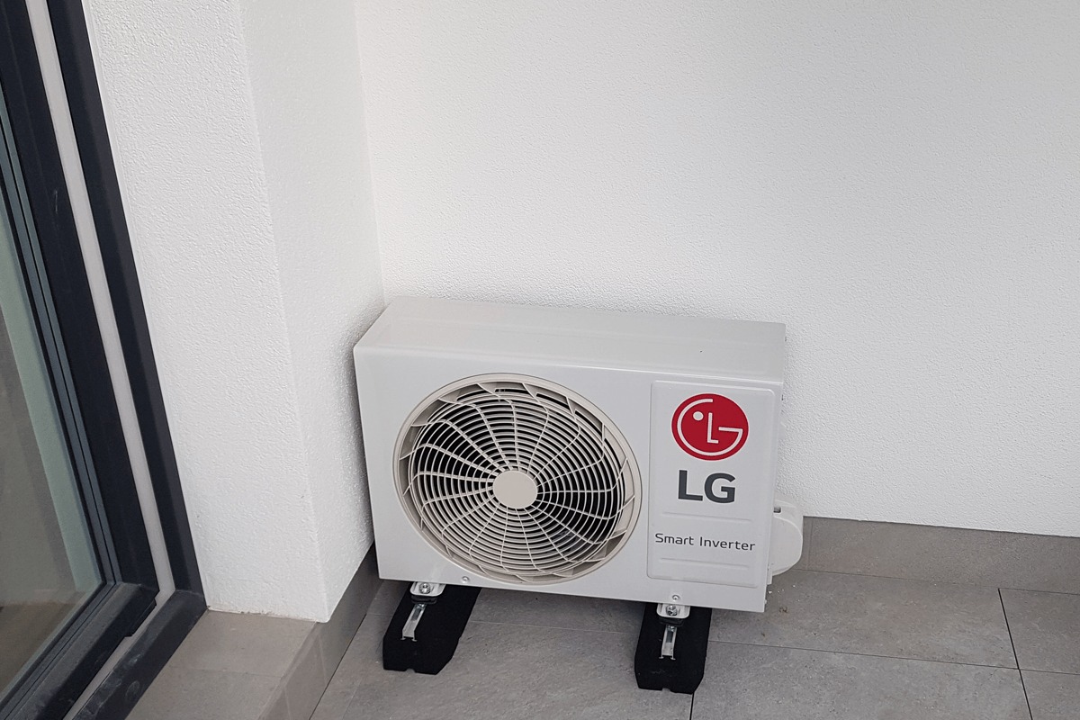Biała zewnętrzna jednostka klimatyzacji LG Smart Inverter zamontowana na czarnych podkładkach w rogu balkonu, widoczna biała ściana i fragment ramy okiennej.