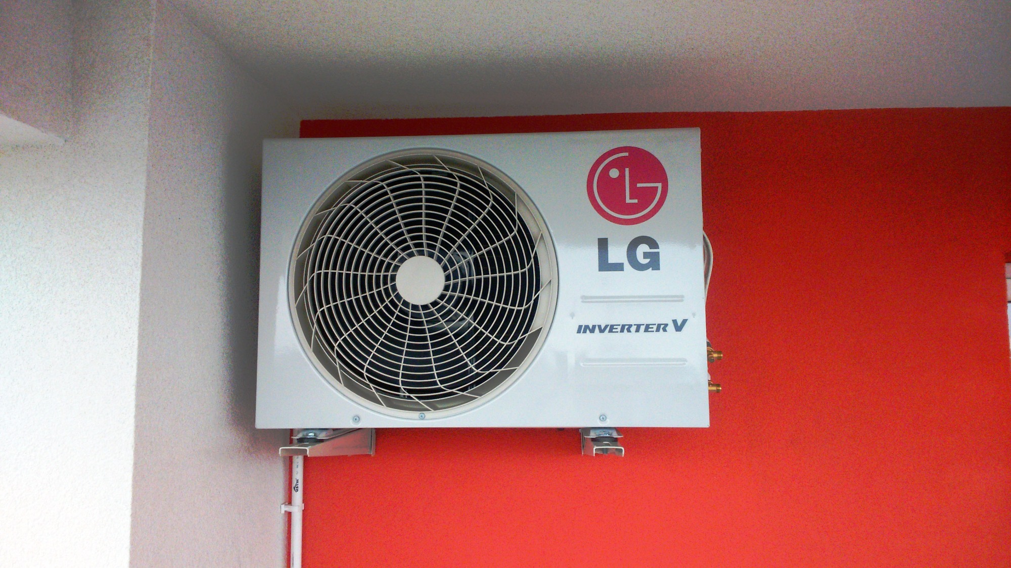 Zewnętrzna jednostka klimatyzacji LG Inverter V zamontowana na czerwonej ścianie, widoczne metalowe wsporniki i rurki.