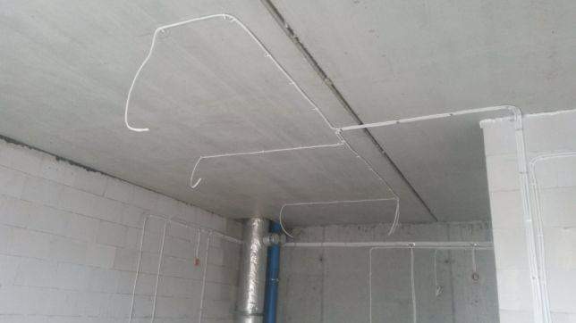 Białe kable elektryczne prowadzone po betonowym suficie i ścianach w surowym wnętrzu, widoczny pionowy przewód wentylacyjny w metalowej izolacji.