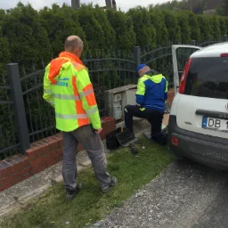 Dwóch mężczyzn w odblaskowych kurtkach pracuje przy skrzynce elektrycznej obok białego samochodu na tle żywopłotu i ogrodzenia.