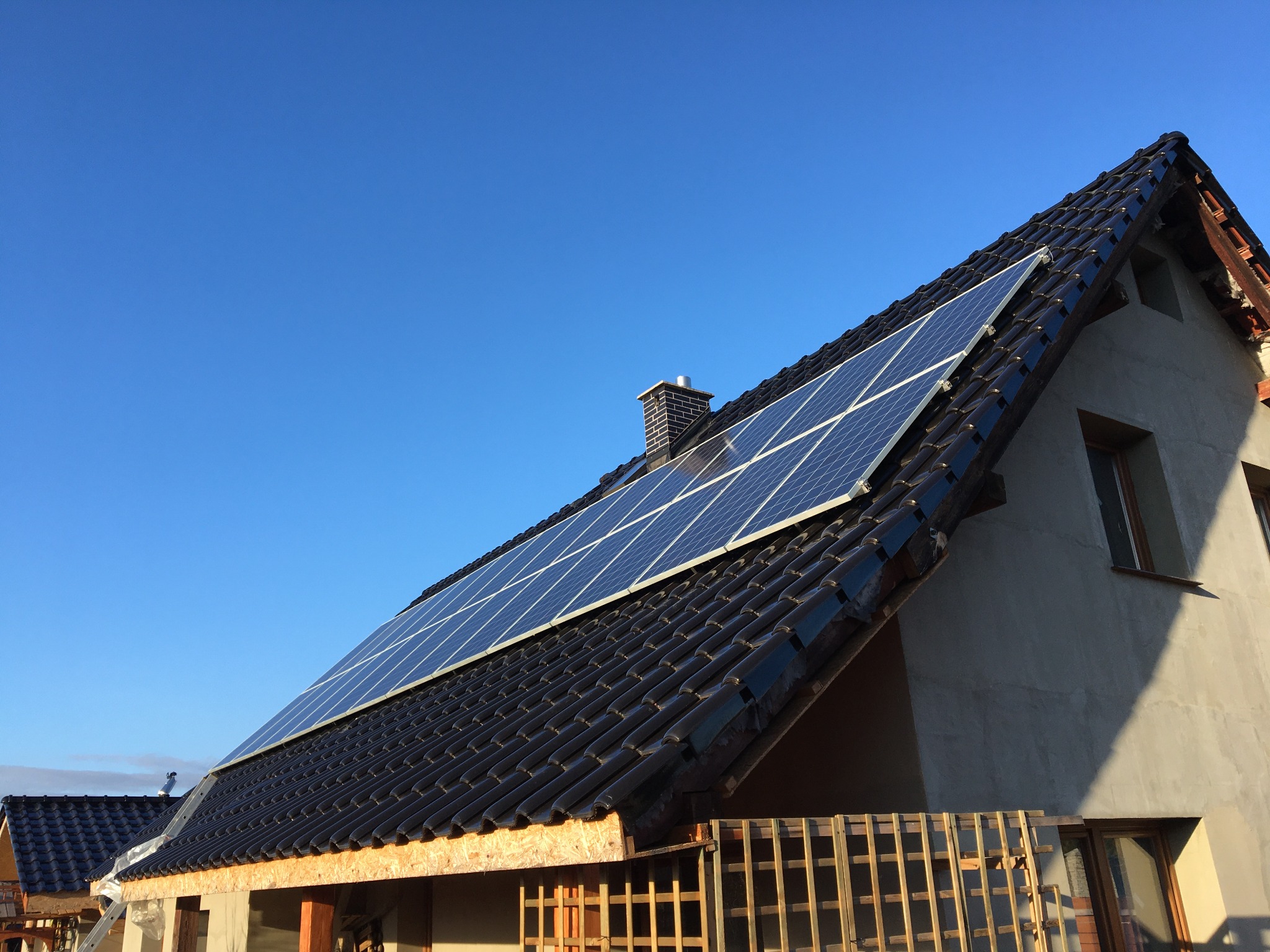 Panele słoneczne zamontowane na dachu domu jednorodzinnego z czarną dachówką, widoczne rusztowania u dołu elewacji i błękitne niebo w tle.