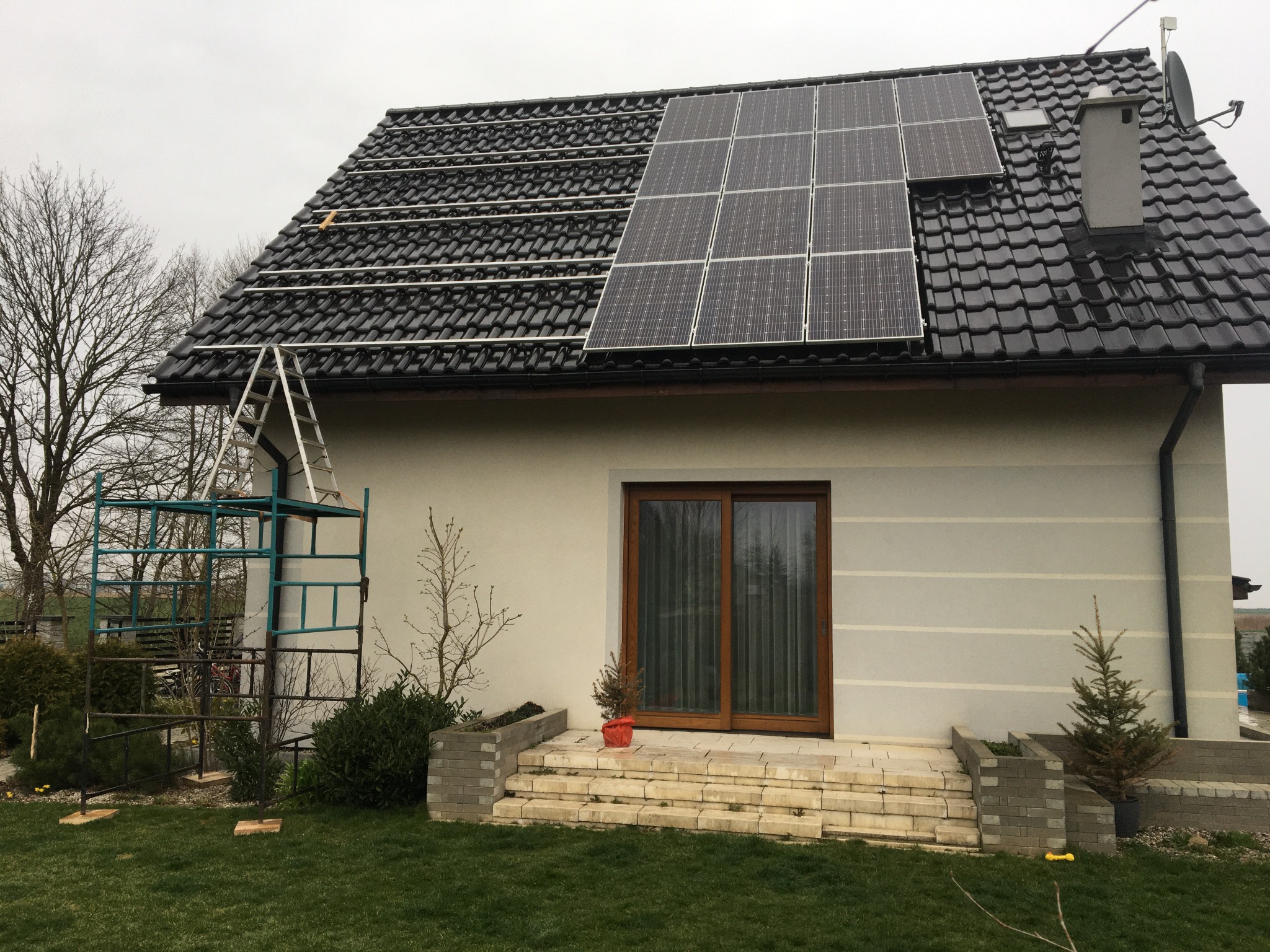 Montaż paneli słonecznych na dachu domu jednorodzinnego z czarną dachówką, widoczne rusztowanie i drabina, część paneli już zamontowana.