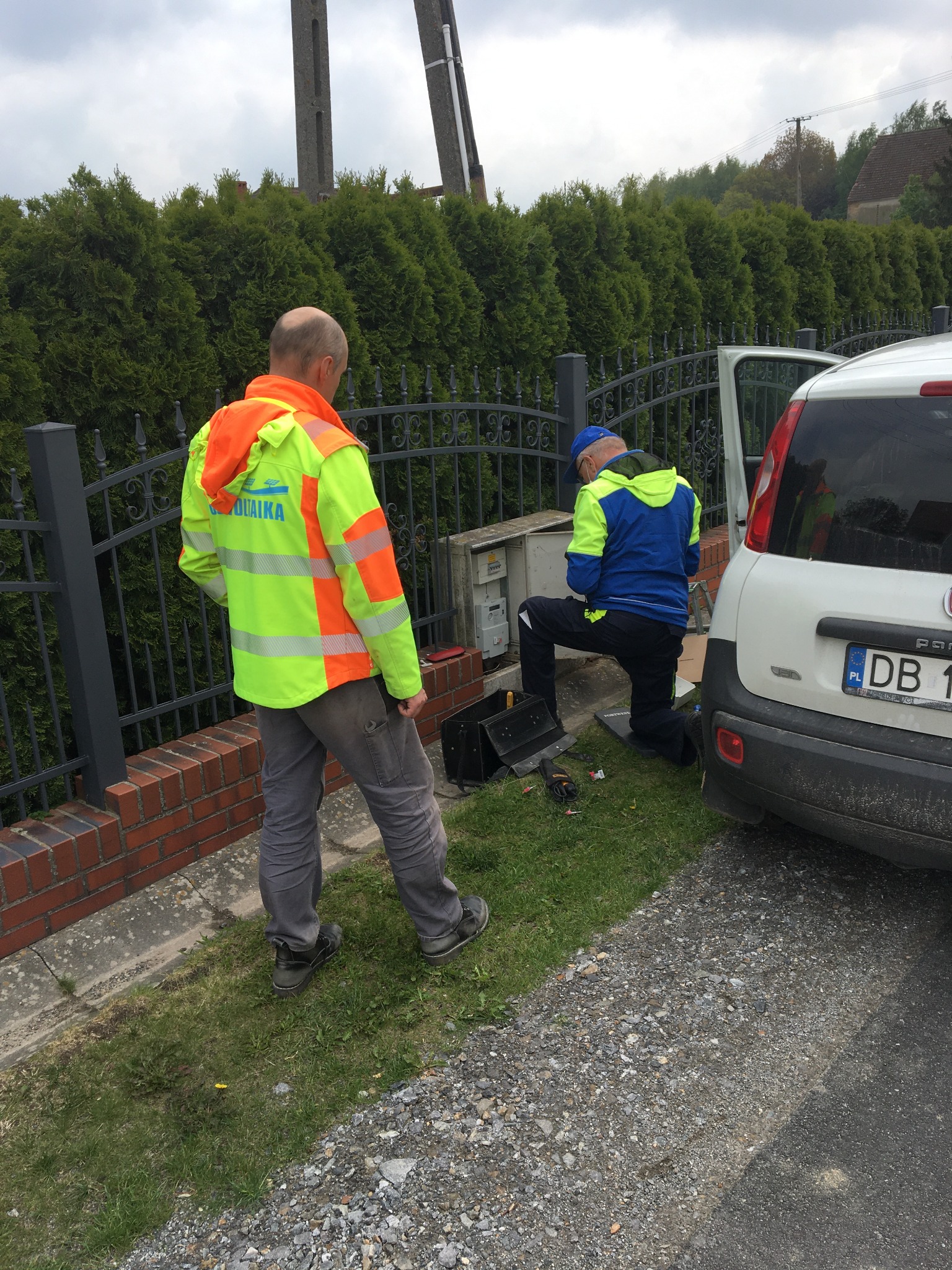 Dwóch mężczyzn w odblaskowych kurtkach pracuje przy skrzynce elektrycznej obok białego samochodu na tle żywopłotu i ogrodzenia.
