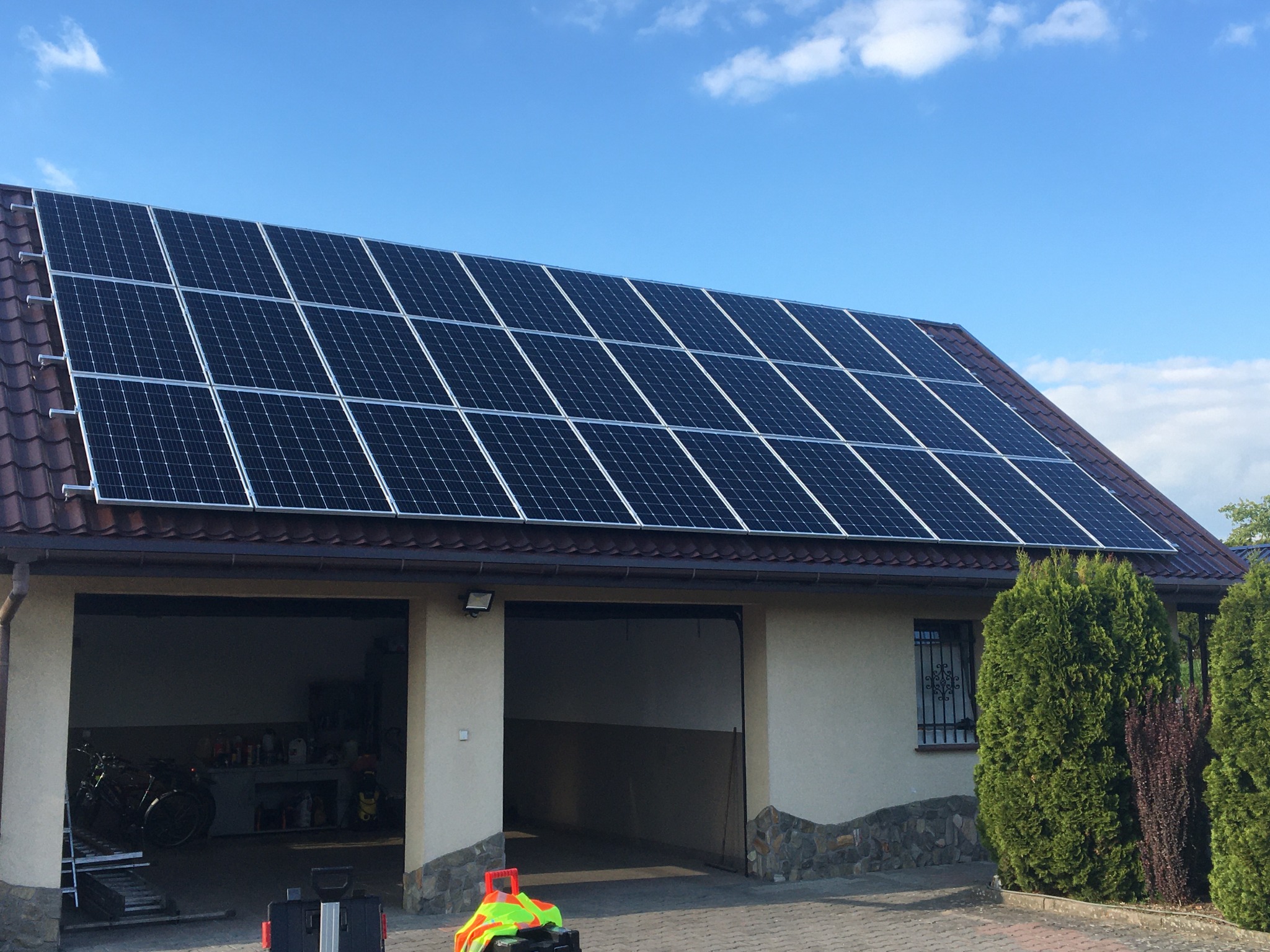 Panele słoneczne zamontowane na dachu domu jednorodzinnego z dwoma garażami, widoczne narzędzia i akcesoria na podjeździe.