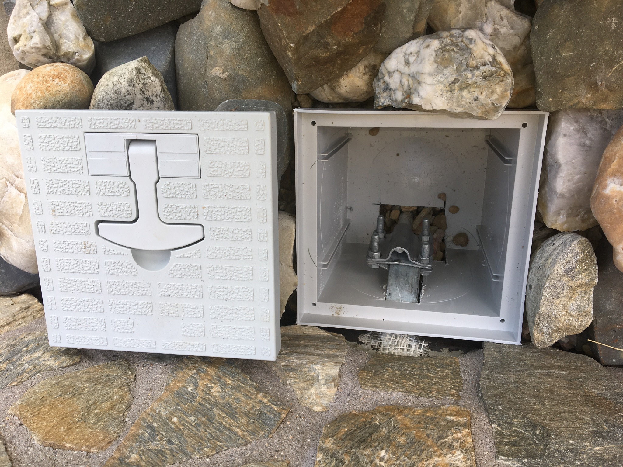 Otwarta, biała skrzynka instalacyjna z metalowymi elementami wewnątrz, umieszczona na kamiennej posadzce i otoczona kamieniami różnej wielkości.