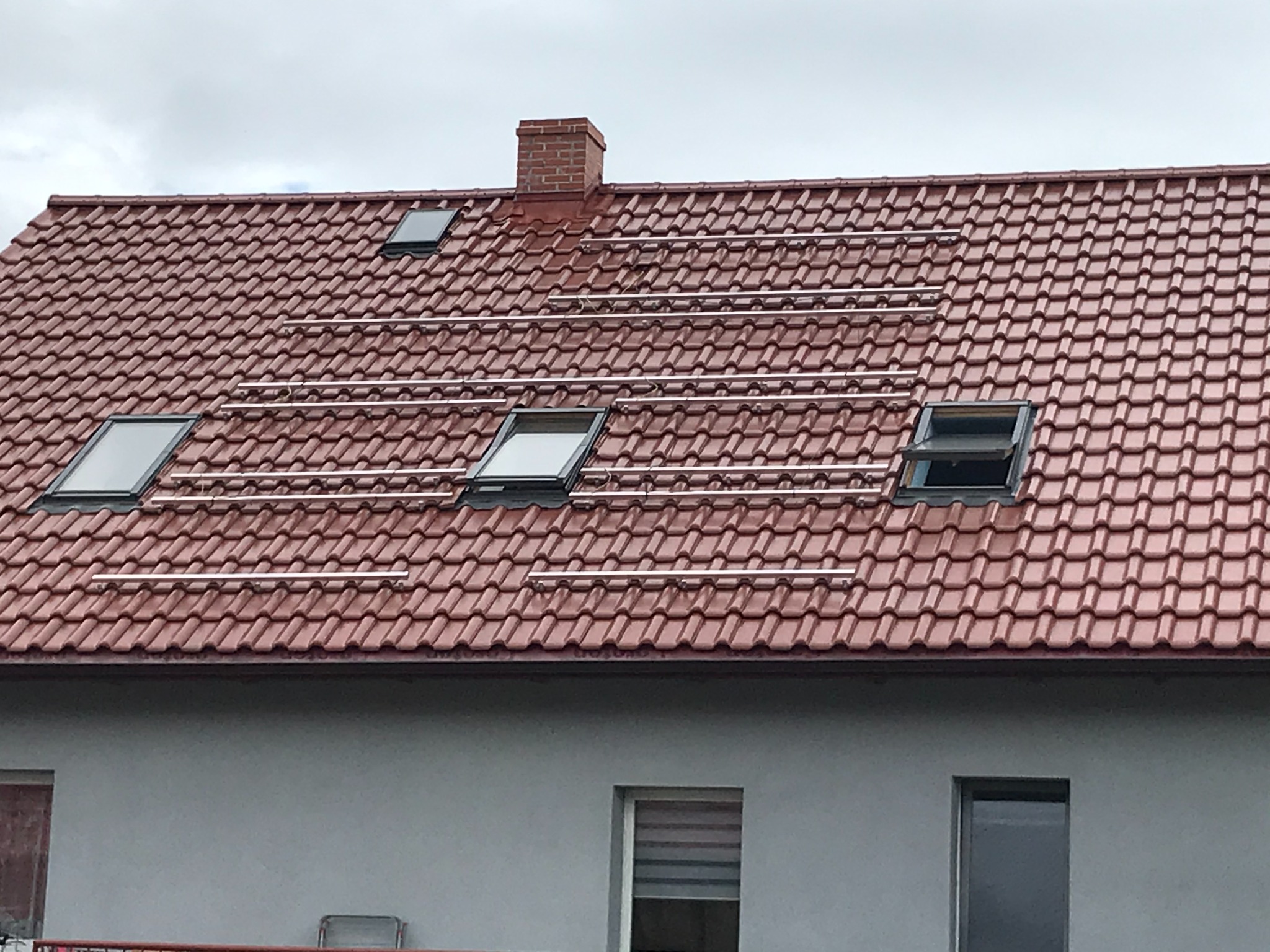 Czerwony dach z dachówkami, na którym zamontowane są szyny montażowe pod panele słoneczne, widoczne okna dachowe i komin.