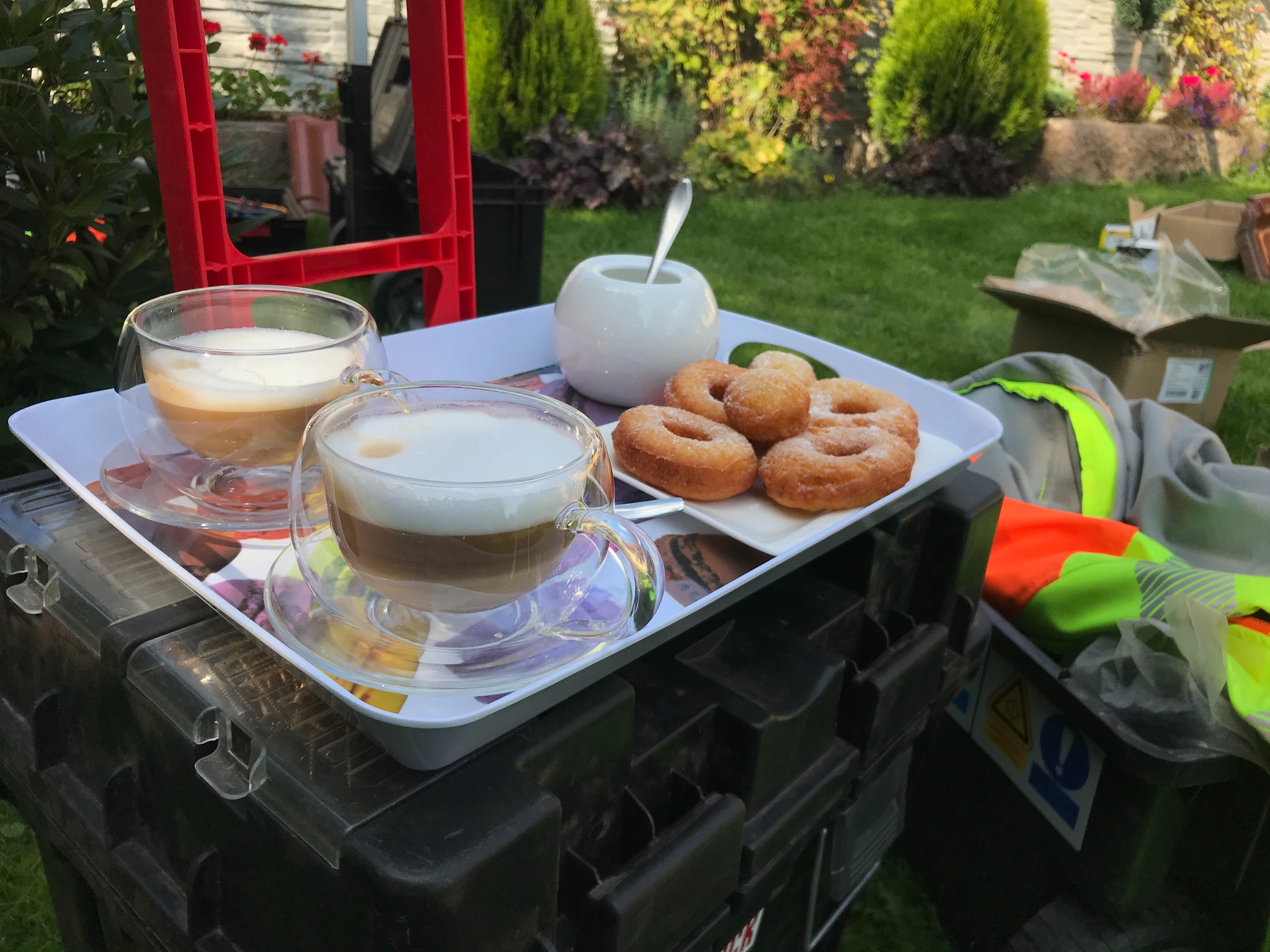Przerwa na kawę i pączki na budowie: Dwie szklanki latte i talerz pączków na tacy, ustawionej na czarnej skrzynce narzędziowej, obok leżącej odblaskowej kamizelki i otwartych kartonów na trawniku...