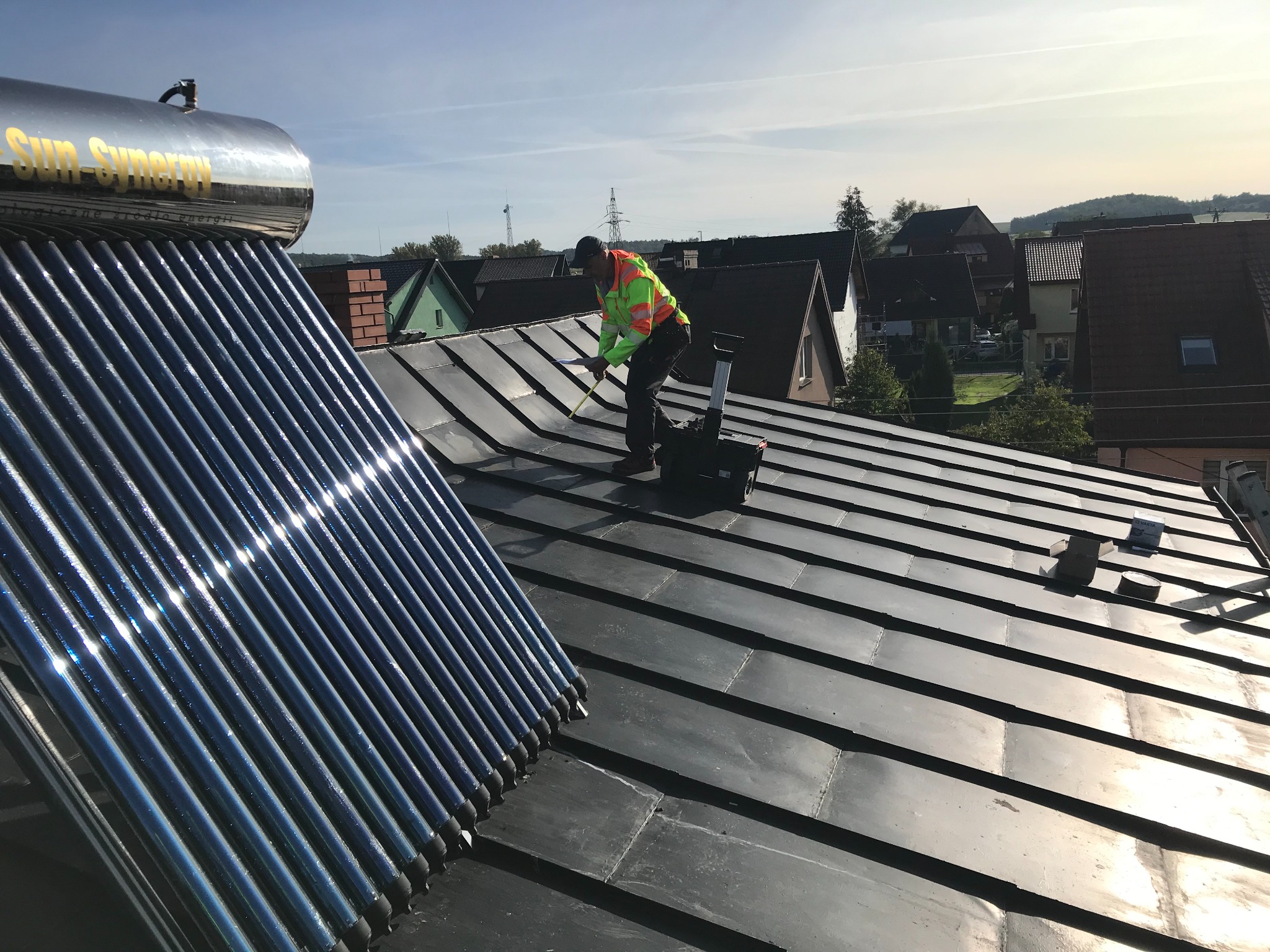 Montaż kolektorów słonecznych na dachu z blachy, pracownik w odblaskowej kamizelce mierzy odległość, w tle zabudowa jednorodzinna i błękitne niebo.