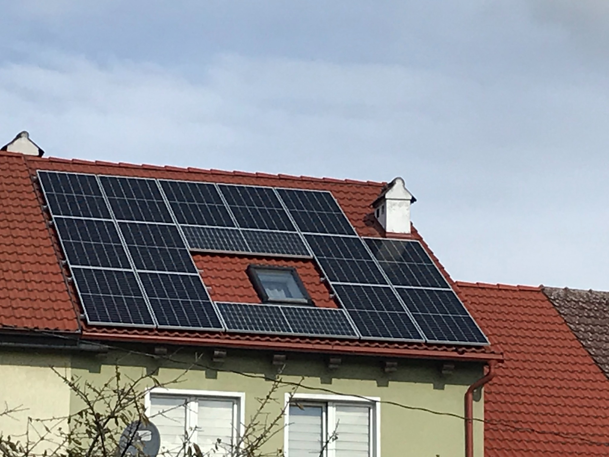 Panele słoneczne na dachu z czerwoną dachówką, z oknem dachowym pośrodku, widok z dołu na tle pochmurnego nieba.