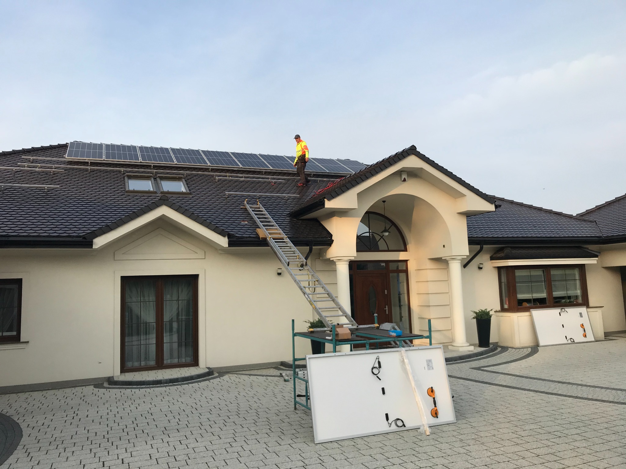 Montaż paneli słonecznych na dachu domu w Ziębicach. Pracownik w odblaskowej kamizelce na dachu, panele częściowo zamontowane, drabina oparta o dach, panele na podwórku na stojaku.