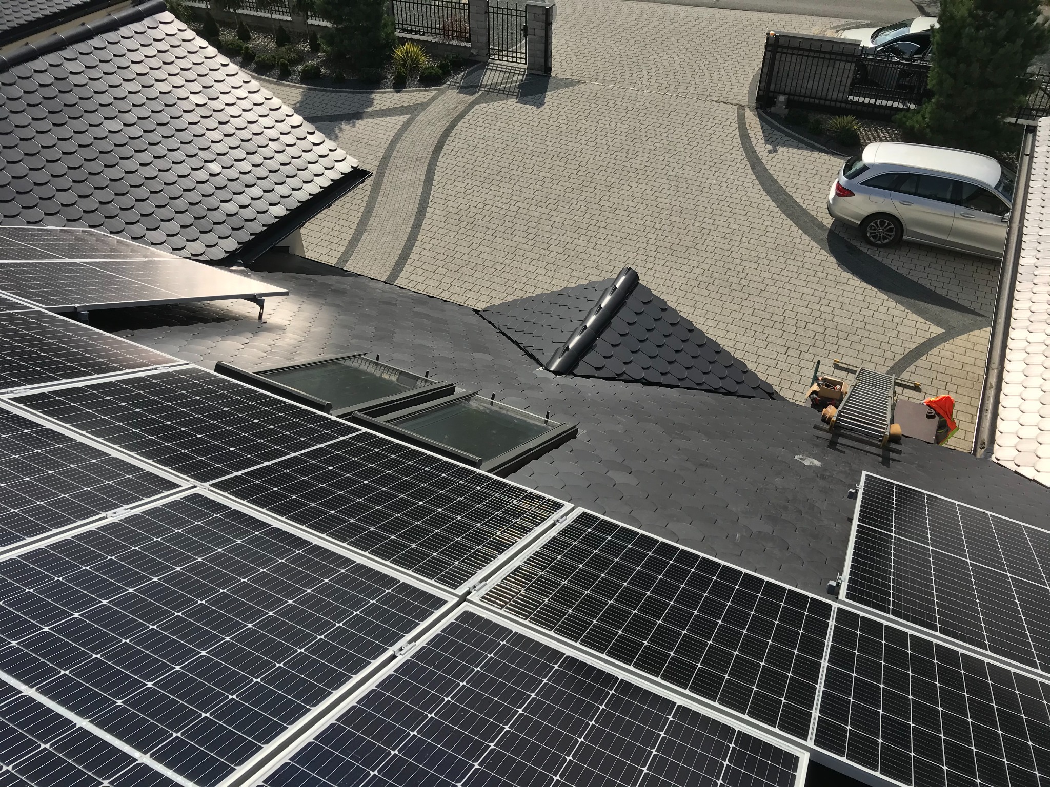 Widok z góry na dach z panelami fotowoltaicznymi i oknami dachowymi na tle podjazdu z kostki brukowej i zaparkowanego samochodu.