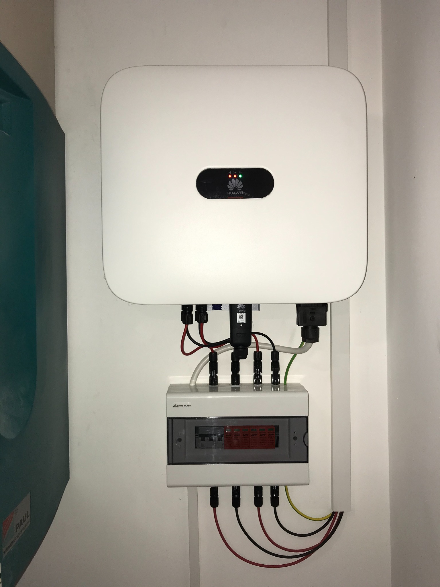 Biały inwerter Huawei z podłączonymi kablami i rozdzielnicą elektryczną na białej ścianie, widoczne diody sygnalizacyjne.