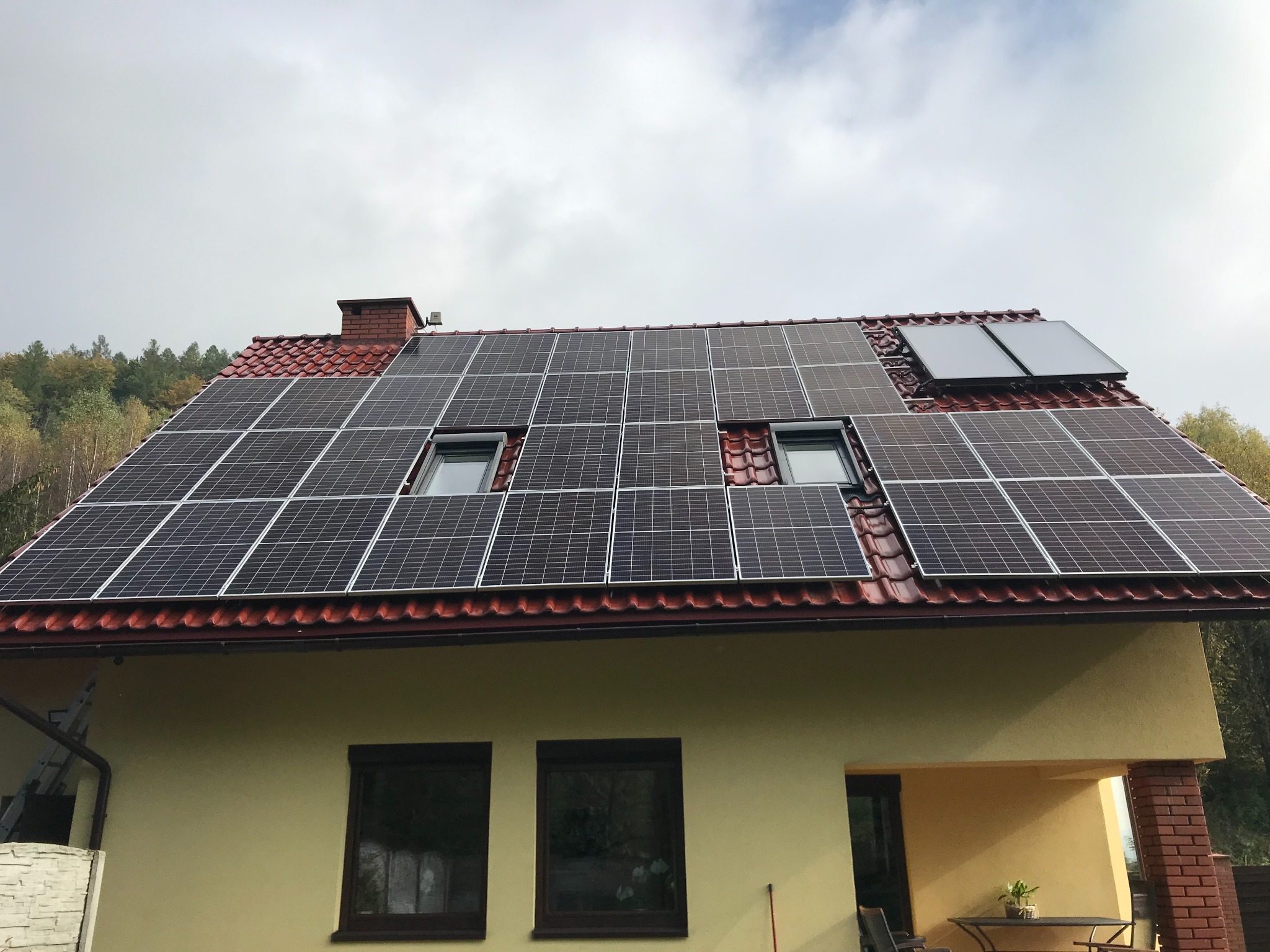 Dom z panelami słonecznymi na dachu z czerwonej dachówki, widok na zielone drzewa w tle i pochmurne niebo.