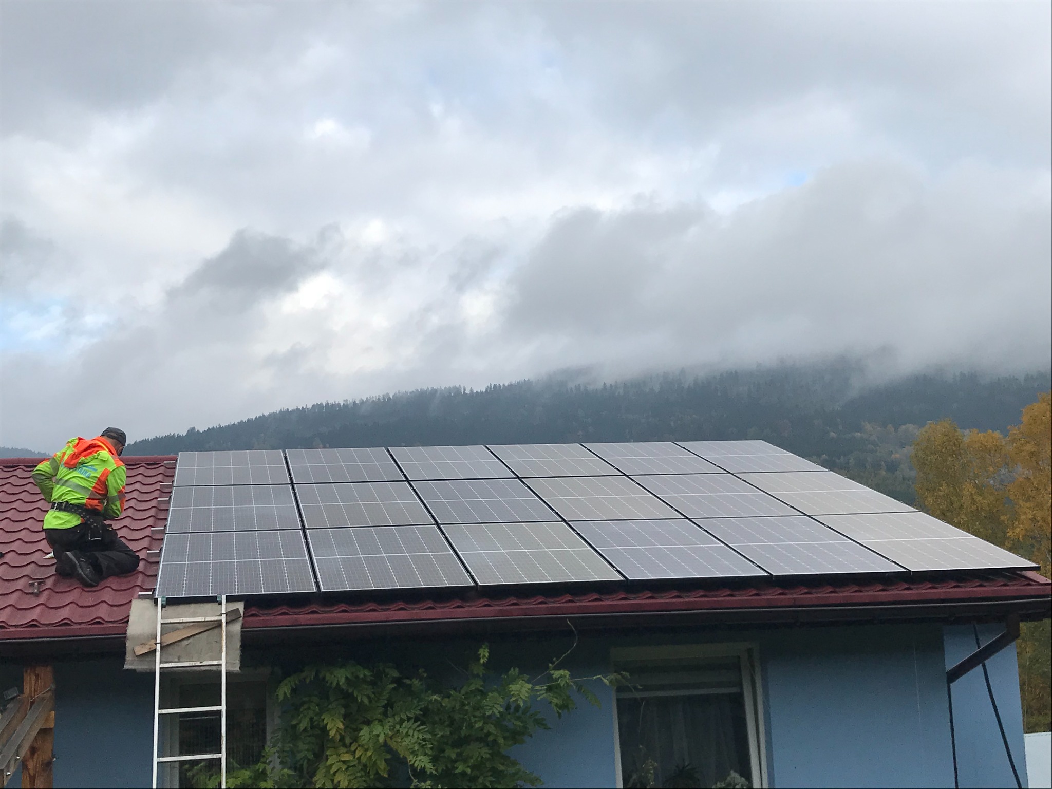 Instalator w odblaskowej kamizelce montuje panele słoneczne na dachu domu z czerwoną dachówką; w tle zalesione wzgórza spowite mgłą.