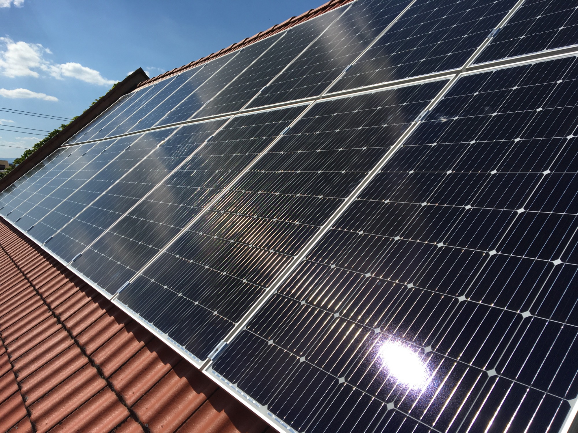 Panele słoneczne zamontowane na dachu z czerwonej dachówki, odbijające promienie słoneczne i błękitne niebo.