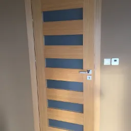 Jasne, drewniane drzwi wewnętrzne z przeszkleniami w poziomych pasach, widoczne na tle beżowej ściany z włącznikami i termostatem.