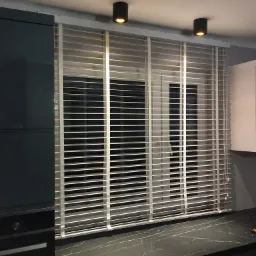 Wnętrze kuchni z białymi żaluzjami poziomymi w oknie, widoczne czarne lampy sufitowe, ciemnoszara zabudowa kuchenna i fragment blatu z narzędziem.