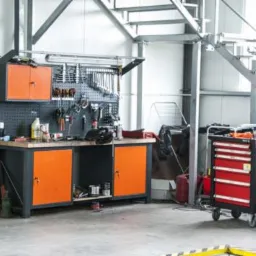 Warsztat samochodowy: stół roboczy z narzędziami na ścianie, czerwona szafka narzędziowa na kółkach, schody metalowe w tle.