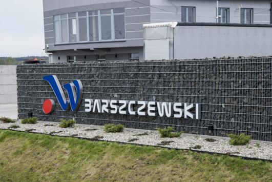 Tablica z logo i nazwą firmy 'Barszczewski' na tle gabionowego muru, przed budynkiem biurowym.