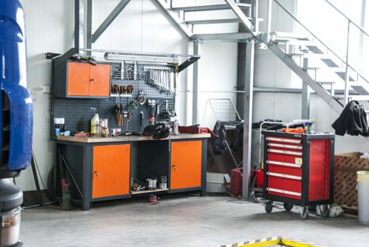 Warsztat samochodowy: stół roboczy z narzędziami na ścianie, czerwona szafka narzędziowa na kółkach, schody metalowe w tle.