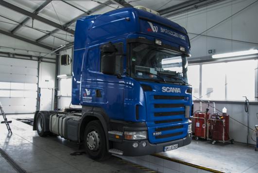 Niebieski ciągnik siodłowy marki Scania R420, z widocznym logo firmy transportowej 'Barszczewski', zaparkowany w warsztacie na kanale inspekcyjnym.