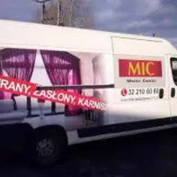 Biały samochód dostawczy z reklamą okien i dekoracji okiennych, w tym fioletowych zasłon i karniszy. Na boku pojazdu widoczne logo firmy MIC oraz numer telefonu. Reklama naklejona na karoserii.