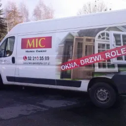 Biały samochód dostawczy z reklamą firmy MIC: okna, drzwi, rolety, numer telefonu i adres strony internetowej.
