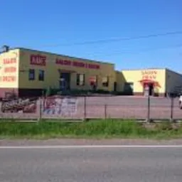 Budynek salonu sprzedaży stolarki okiennej i drzwiowej w kolorze żółtym z czerwonymi napisami, ogrodzony metalowym płotem, widok z zewnątrz w słoneczny dzień.