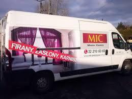 Biały samochód dostawczy z reklamą okien i dekoracji okiennych, w tym fioletowych zasłon i karniszy. Na boku pojazdu widoczne logo firmy MIC oraz numer telefonu. Reklama naklejona na karoserii.