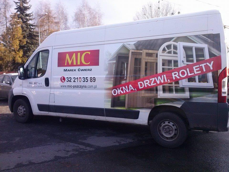 Biały samochód dostawczy z reklamą firmy MIC: okna, drzwi, rolety, numer telefonu i adres strony internetowej.