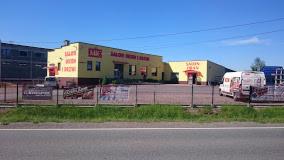 Budynek salonu sprzedaży stolarki okiennej i drzwiowej w kolorze żółtym z czerwonymi napisami, ogrodzony metalowym płotem, widok z zewnątrz w słoneczny dzień.