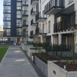 Nowoczesny budynek wielorodzinny z balkonami i przeszkloną fasadą, widok z poziomu chodnika i trawnika w słoneczny dzień.