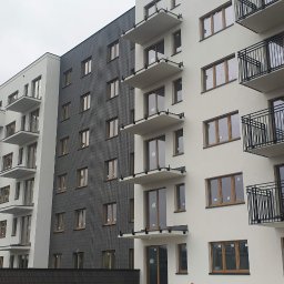 Macrotech Sp z o.o. Sp. Komandytowa - Nowoczesny budynek wielorodzinny z elewacją w kolorze białym i grafitowym, balkony z czarnymi balustradami, okna z drewnianą stolarką, widok z dołu na kilka kondygnacji.