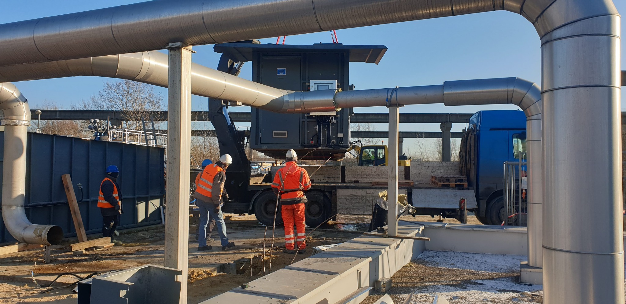 Montaż rurociągu przemysłowego z użyciem dźwigu, pracownicy w odblaskowych kamizelkach nadzorują prace na placu budowy.