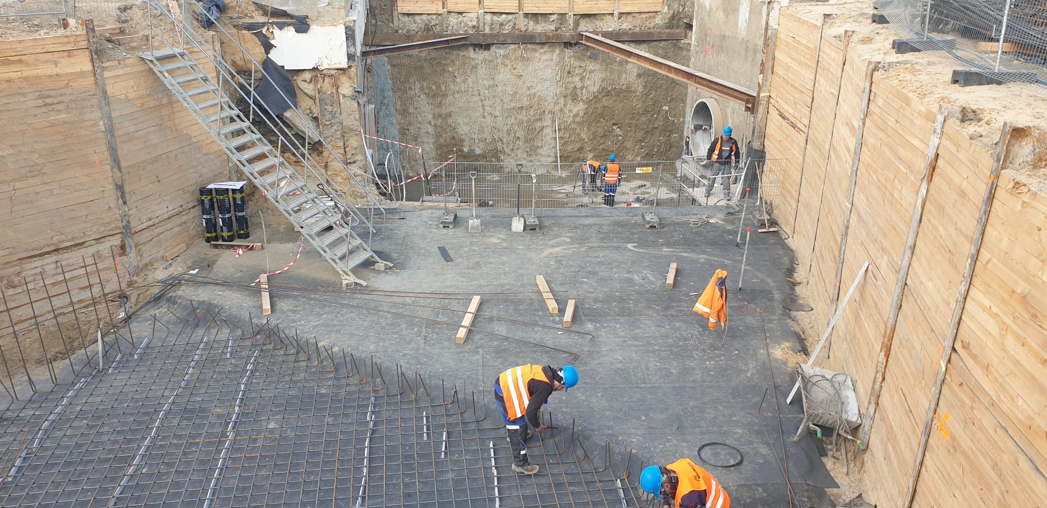 Budowa fundamentów: pracownicy w kaskach i kamizelkach układają zbrojenie na czarnej folii w wykopie zabezpieczonym drewnianą szalunką, widoczna drabina i materiały izolacyjne.