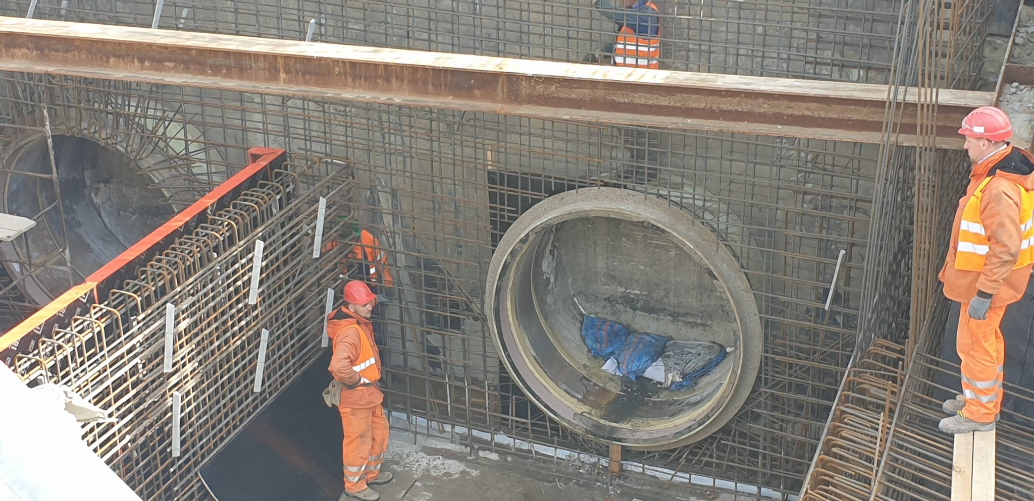 Budowa infrastruktury podziemnej: robotnicy w pomarańczowych kombinezonach pracują przy zbrojeniu i betonowych rurach w wykopie.