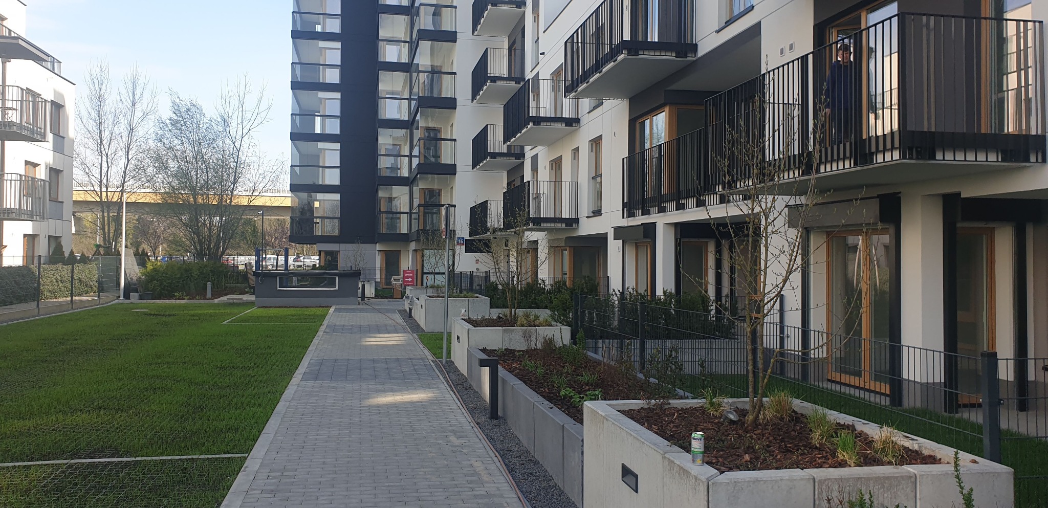 Nowoczesny budynek wielorodzinny z balkonami i przeszkloną fasadą, widok z poziomu chodnika i trawnika w słoneczny dzień.