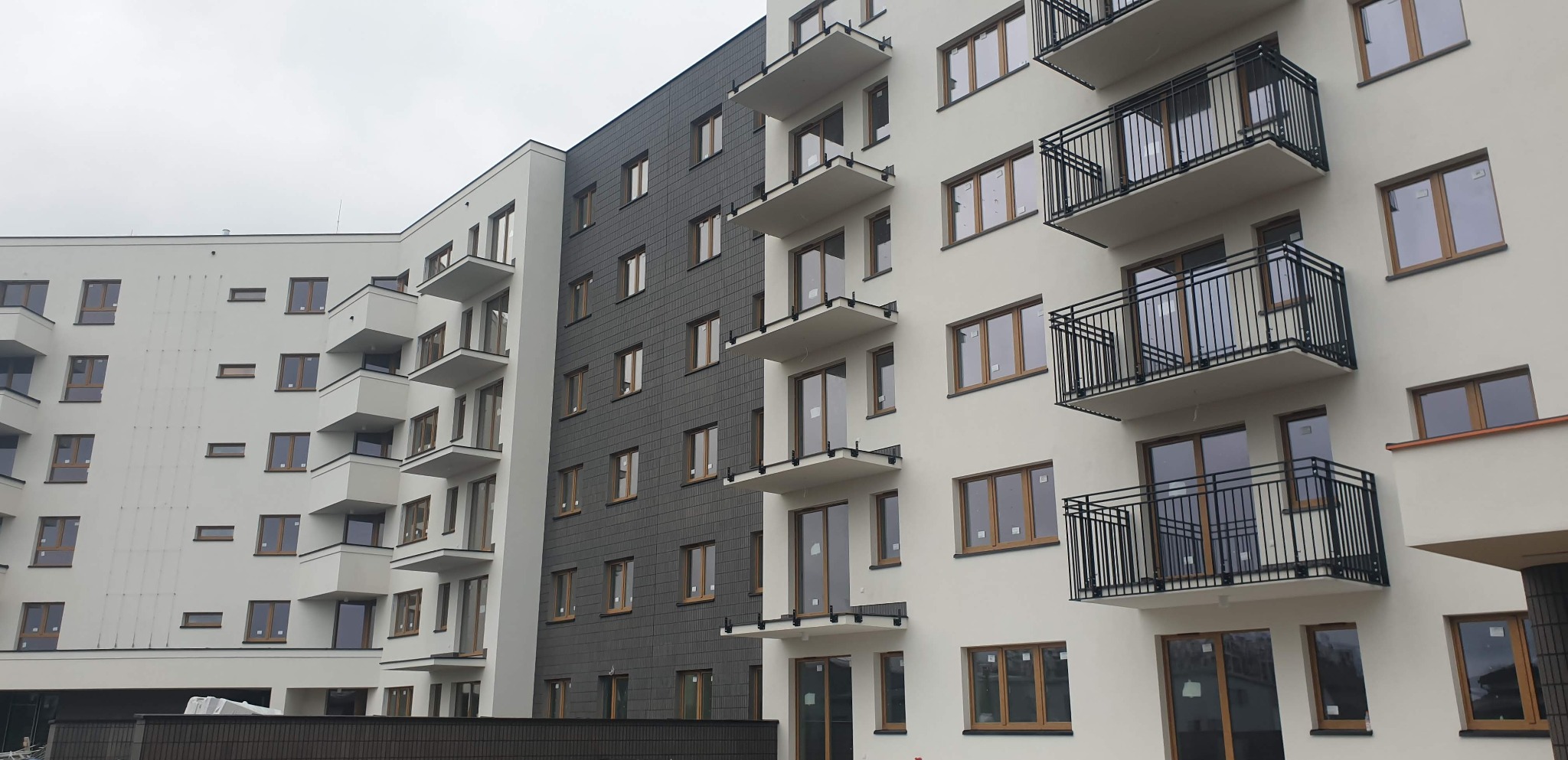 Nowoczesny budynek wielorodzinny z elewacją w kolorze białym i grafitowym, balkony z czarnymi balustradami, okna z drewnianą stolarką, widok z dołu na kilka kondygnacji.