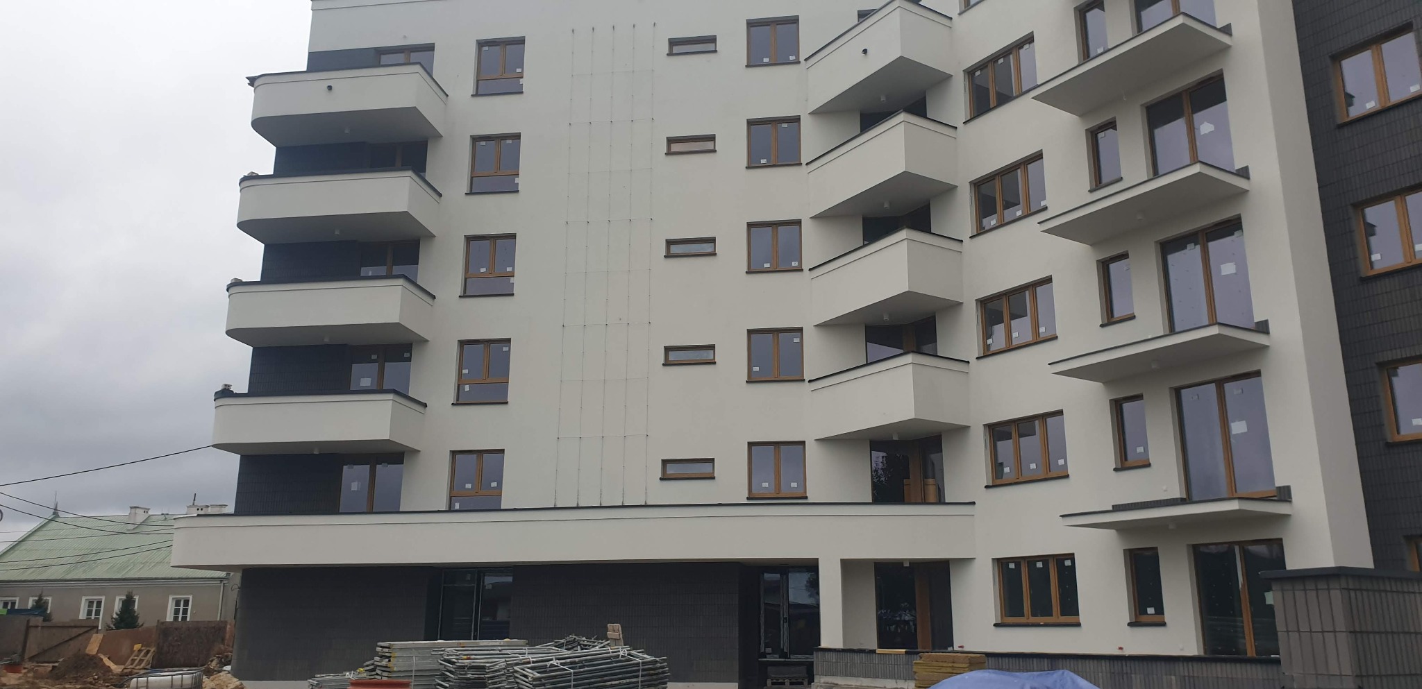 Nowoczesny budynek wielorodzinny z białymi ścianami i brązowymi ramami okiennymi, balkony z czarnymi elementami wykończeniowymi, widoczne rusztowania i materiały budowlane u podstawy.