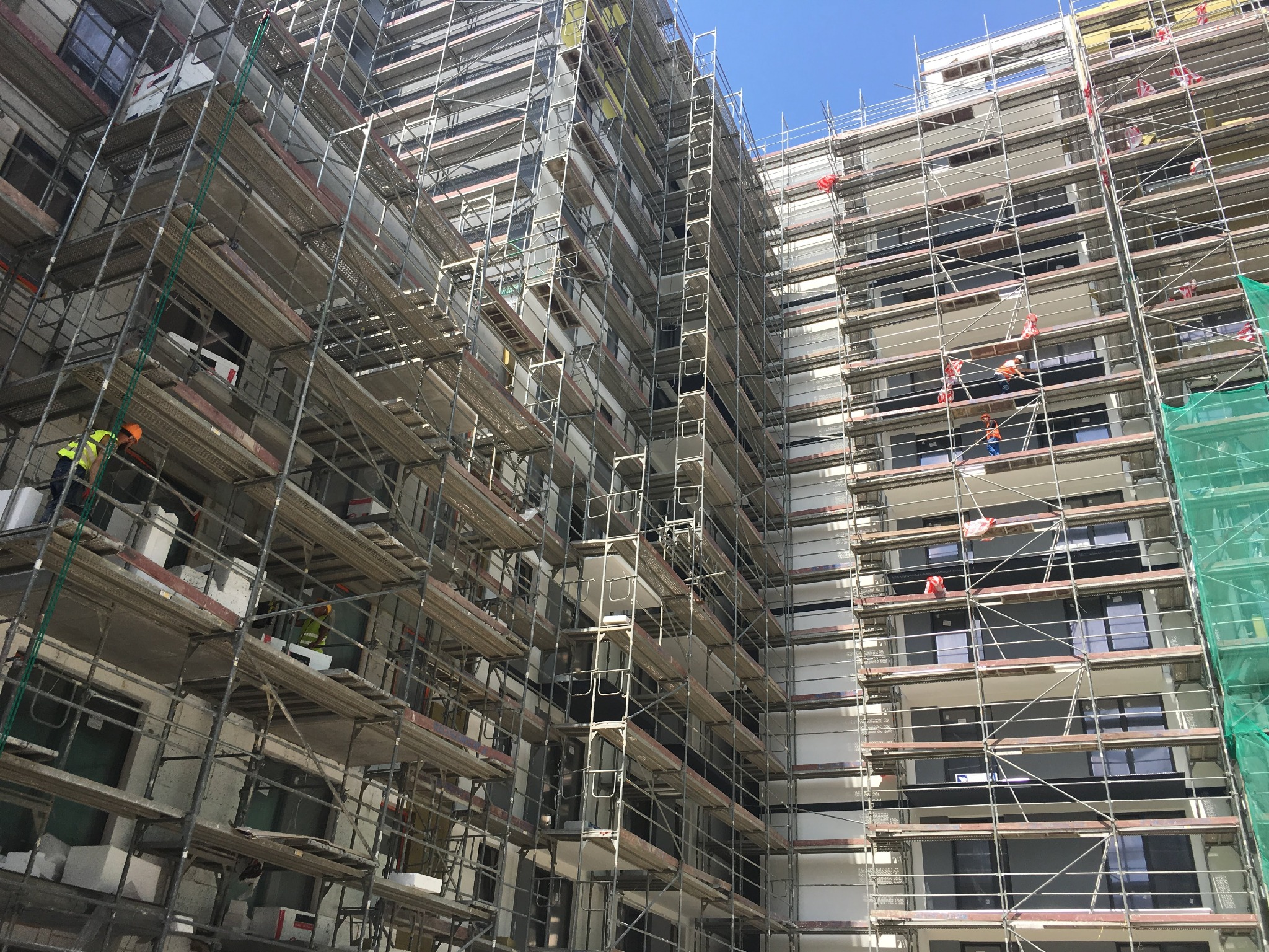 Wielopiętrowy budynek w trakcie prac elewacyjnych, otoczony rusztowaniami, z pracownikami w strojach roboczych na różnych poziomach.