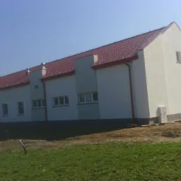 Długi, parterowy budynek z jasną elewacją i czerwoną dachówką, widoczny z perspektywy trawnika, z elementami wykończeniowymi wokół budynku i zaparkowanym samochodem dostawczym w tle.