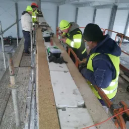 Pracownicy w odblaskowych kamizelkach układają bloczki z betonu komórkowego na ścianie szkieletowej budynku przemysłowego, widoczne rusztowania i narzędzia pracy.