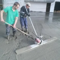 Dwóch pracowników w gumowych butach wyrównuje wylewkę betonową za pomocą listwy wibracyjnej z silnikiem spalinowym i długiego drewnianego narzędzia w surowym stanie budynku.