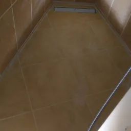 Wykończony brodzik z brązowymi płytkami, odpływ liniowy w rogu, widoczny wąż prysznicowy