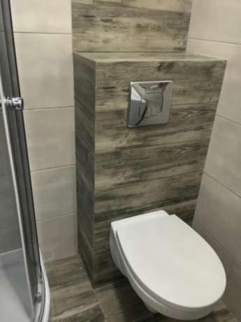 Nowoczesna toaleta podwieszana z zabudową w okleinie drewnopodobnej, chromowany przycisk spłukiwania, widoczny fragment kabiny prysznicowej.