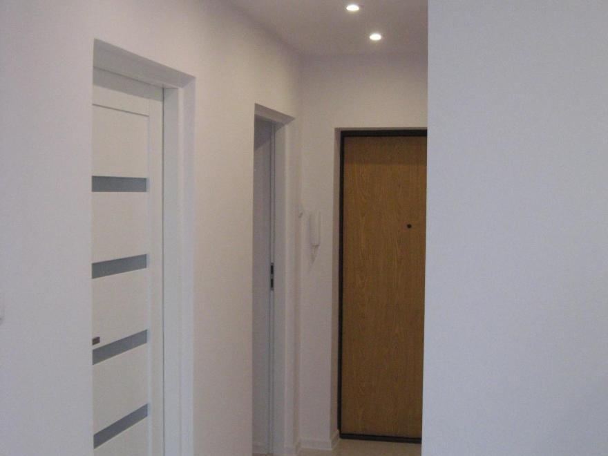 Korytarz z trzema drzwiami: białe drzwi wewnętrzne z czterema poziomymi, szarymi paskami, białe drzwi do pomieszczenia gospodarczego i drewniane drzwi wejściowe z czarną ramą i wizjerem.