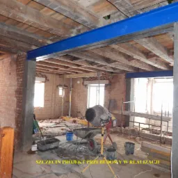 Wnętrze w trakcie przebudowy, z widocznymi stalowymi belkami w kolorze niebieskim, betonowymi filarami, ceglanymi ścianami, drewnianym stropem i betoniarką na środku pomieszczenia.