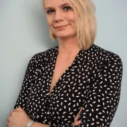 Portret blondynki w czarno-białej sukience z zegarkiem na ręce, stojącej na tle jasnoniebieskiej ściany z założonymi rękami.